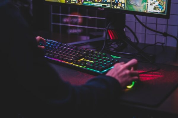 Jemand sitzt vor einem beleuchteten Gaming Rechner mit einer Hand auf der Tastatur und einer auf der Maus
