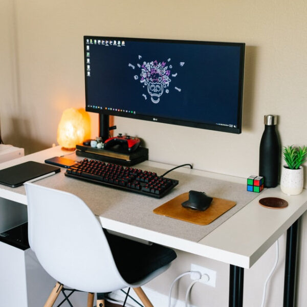 Auf einem weißen Schreibtisch liegt eine beleuchtete Tastatur, Gaming-Maus und ein kleines braunes Mauspad. An der Wandd arüber hängt ein Bildschirm