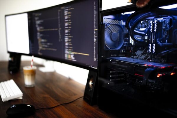 Ein PC mit einem Glasfenster, dessen Komponenten beleuchtet sind. Daneben ein Setup aus zwei Monitoren