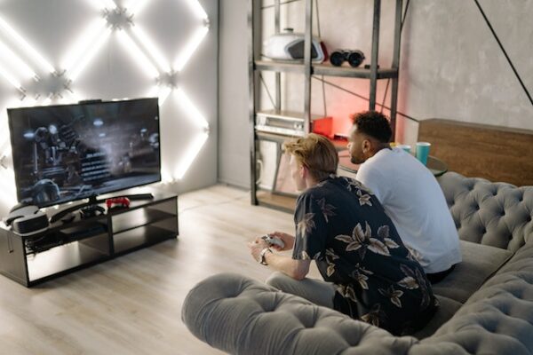 Zwei Männer sitzen auf einer grauen Couch und Spielen ein Spiel am Fernseher, hinter dem LEDs montiert sind