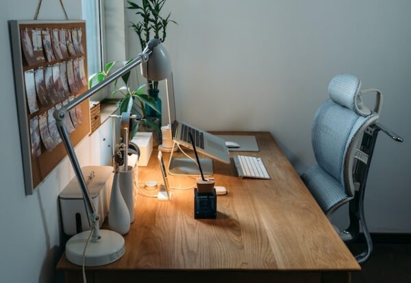 Ein Schreibtisch von der Seite mit Laptopablage, Stehlampe, ergonomischem Stuhl und einer Pinwand voller Bilder an der Wand