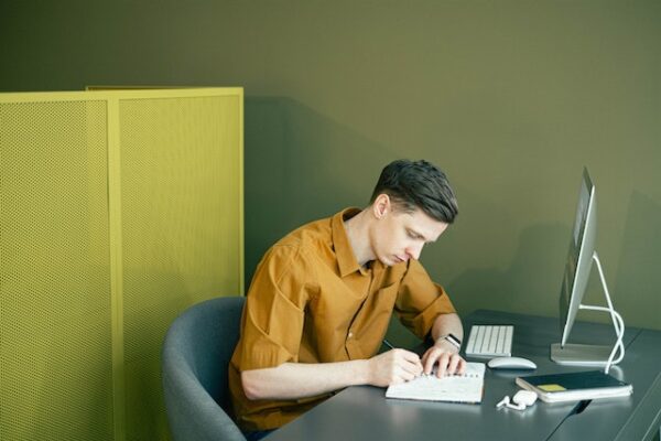 Eine Person sitzt vor Computerbildschirm und Tastatur und schreibt sich Notizen in ein Heft