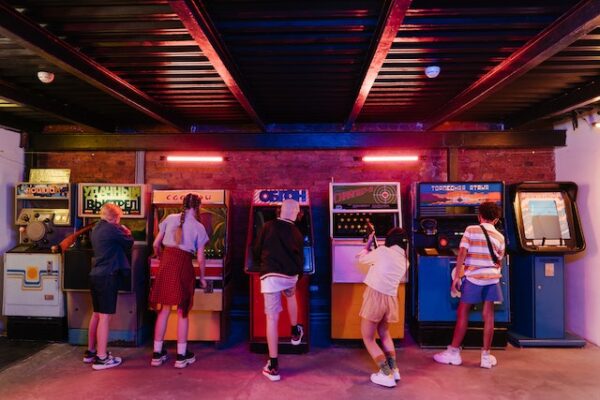 In einer bunt beleuchteten Arcadehalle stehen 5 Personen mit dem Rücken zu Kamera vor Spieleautomaten 
